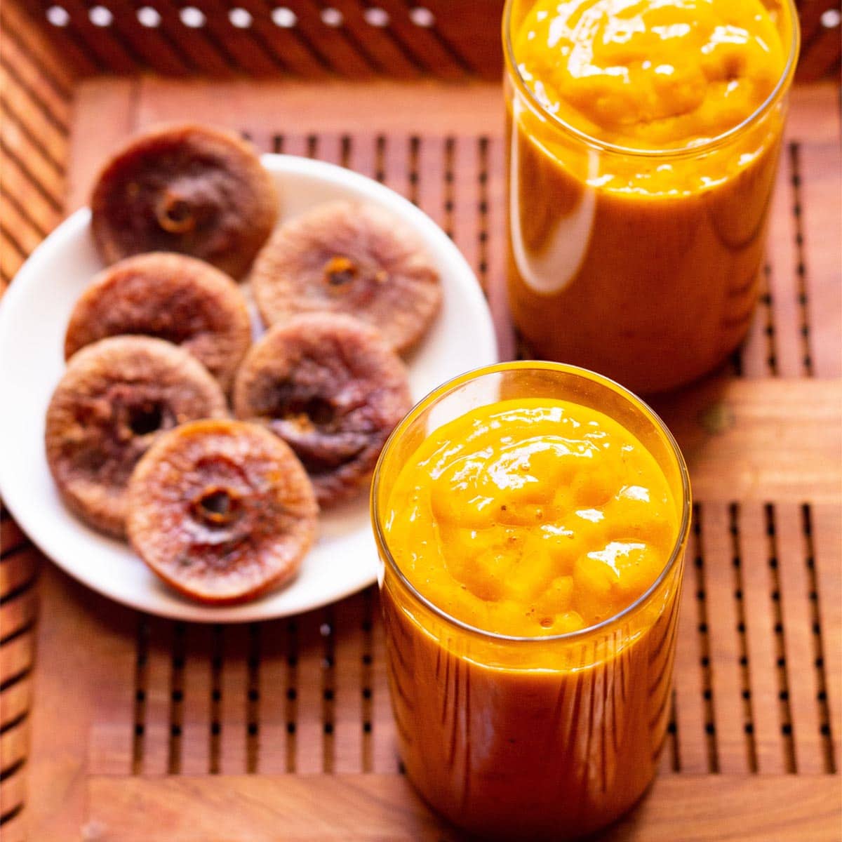 mango smoothie with figs served in two glasses and a white plate of dried figs on a wooden tray