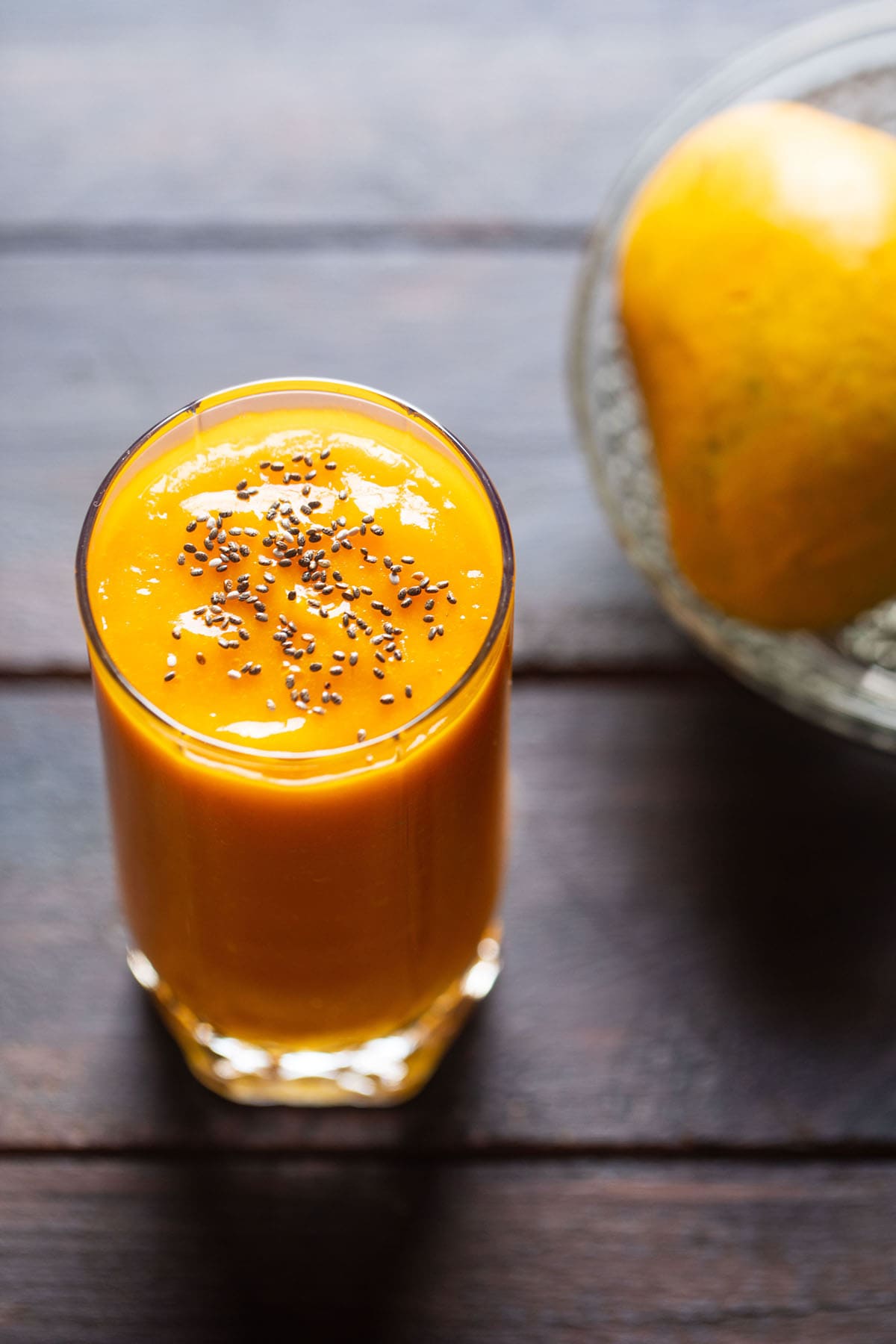 closeup shot mango juice in a glass topped with chia seeds on a wooden table with a side of mangoes on a glass bowl