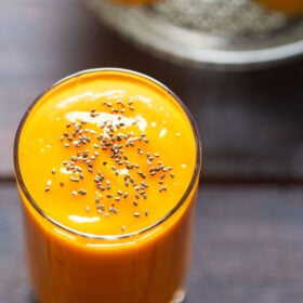 top shot shot of mango juice in a glass topped with chia seeds on a wooden table with mangoes on a glass bowl placed on top