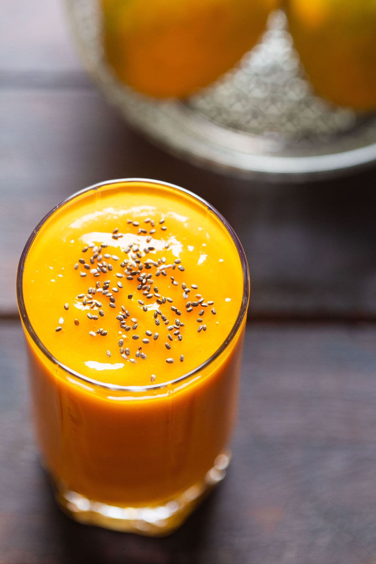 top shot shot of mango juice in a glass topped with chia seeds on a wooden table with mangoes on a glass bowl placed on top