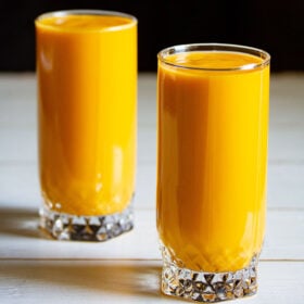 mango milkshake in two tall glasses on white table with a black background