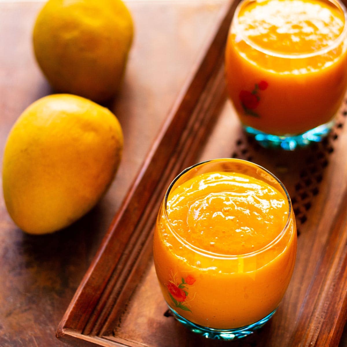 mango papaya banana smoothie served in two small glasses on a wooden tray with two ripe mangoes placed at the side