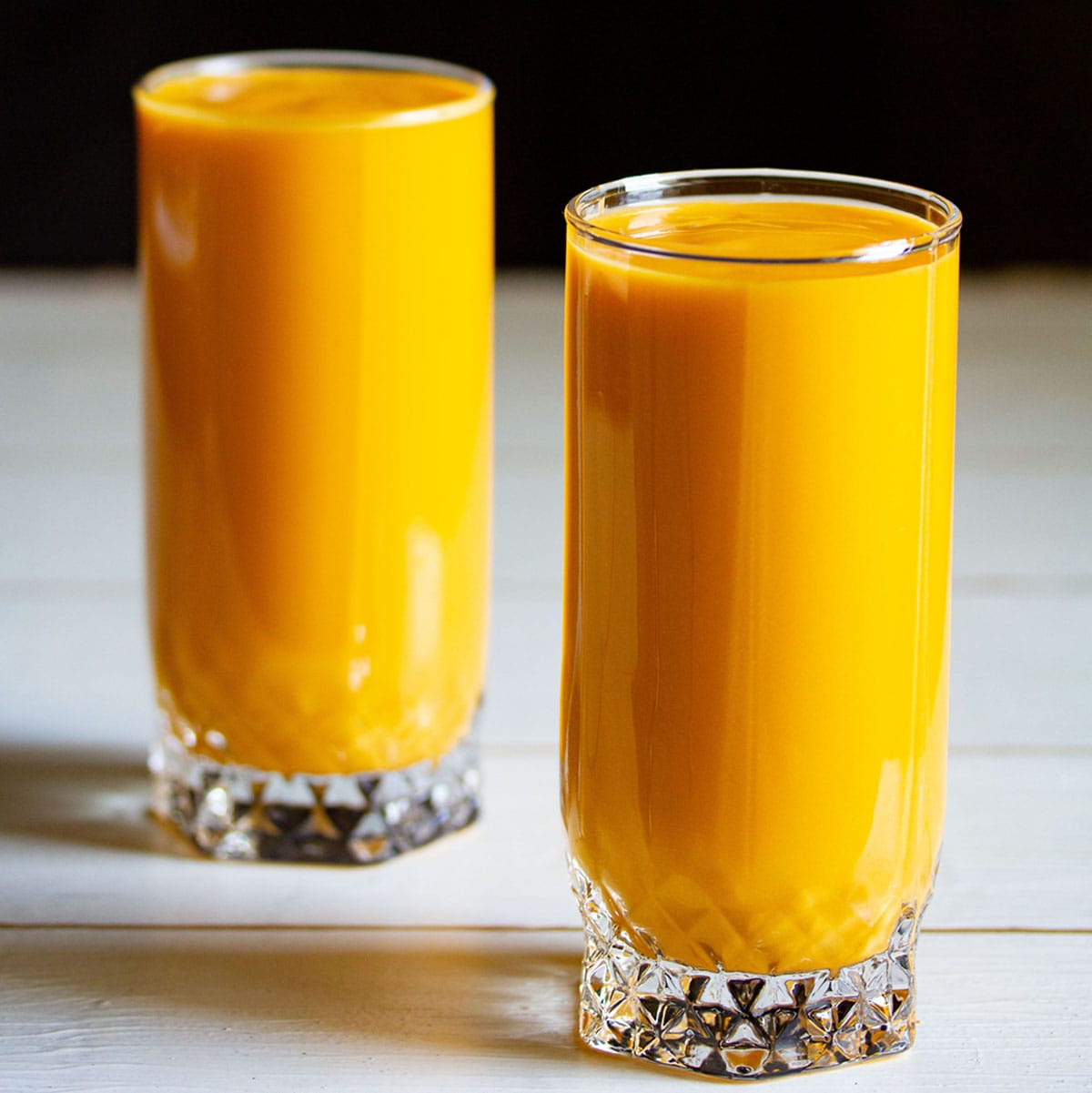 mango milkshake in two tall glasses on white table with a black background 