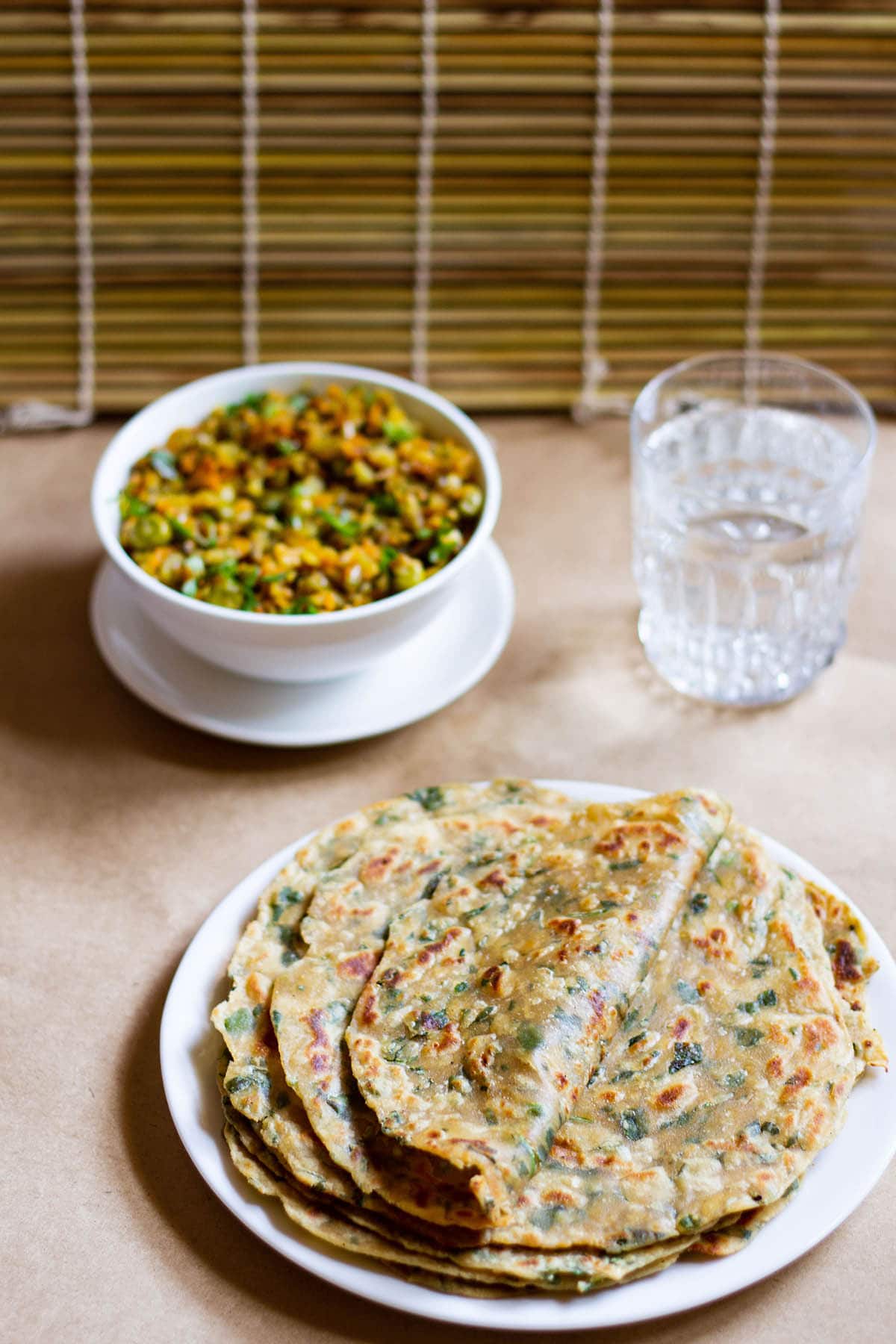 folded methi paratha on a stack of methi parathas in a white plate with a veggie dish in white bowl and glass of water