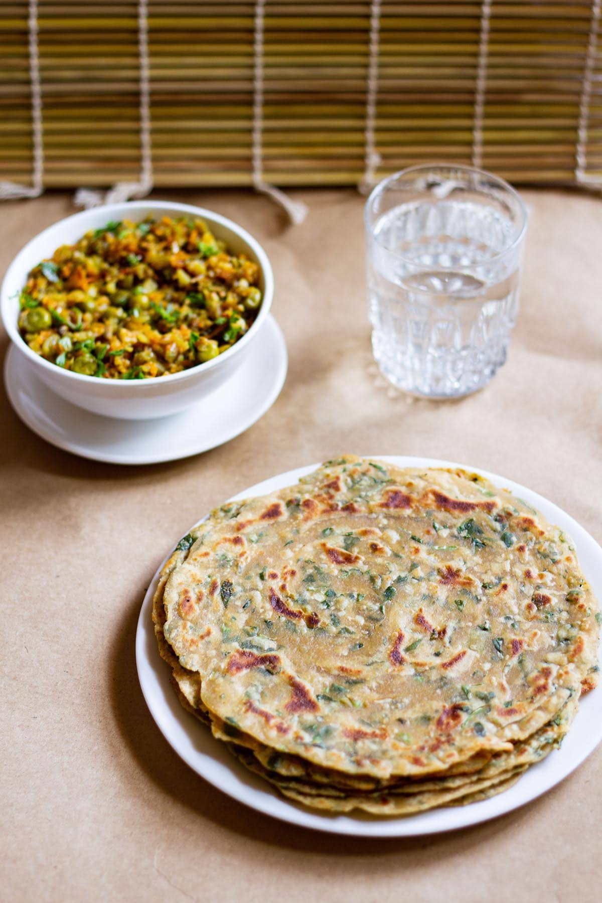 a stack of methi paratha in a white plate with a veggie dish in white bowl and glass of water