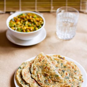 folded methi paratha on a stack of methi parathas in a white plate with a veggie dish in white bowl and glass of water