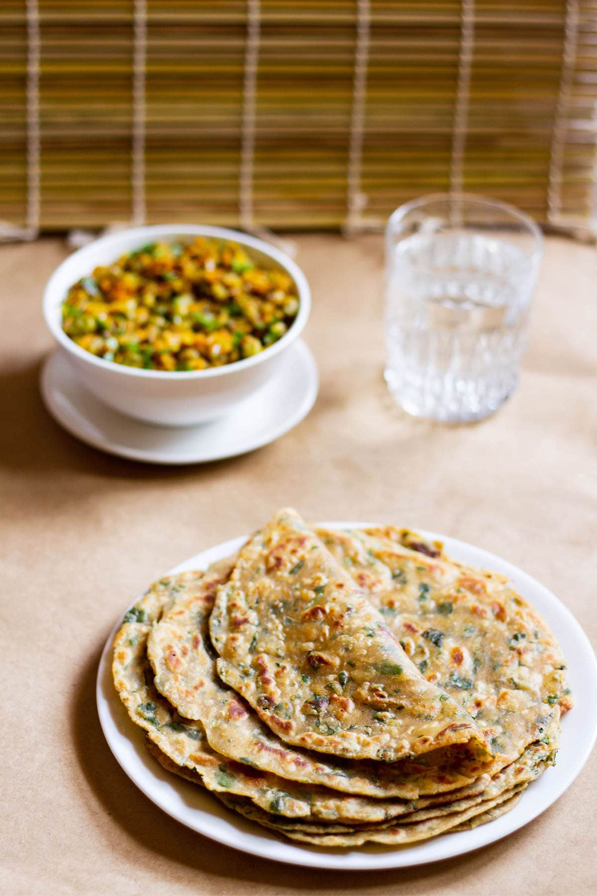 folded methi paratha on a stack of methi parathas in a white plate with a veggie dish in white bowl and glass of water