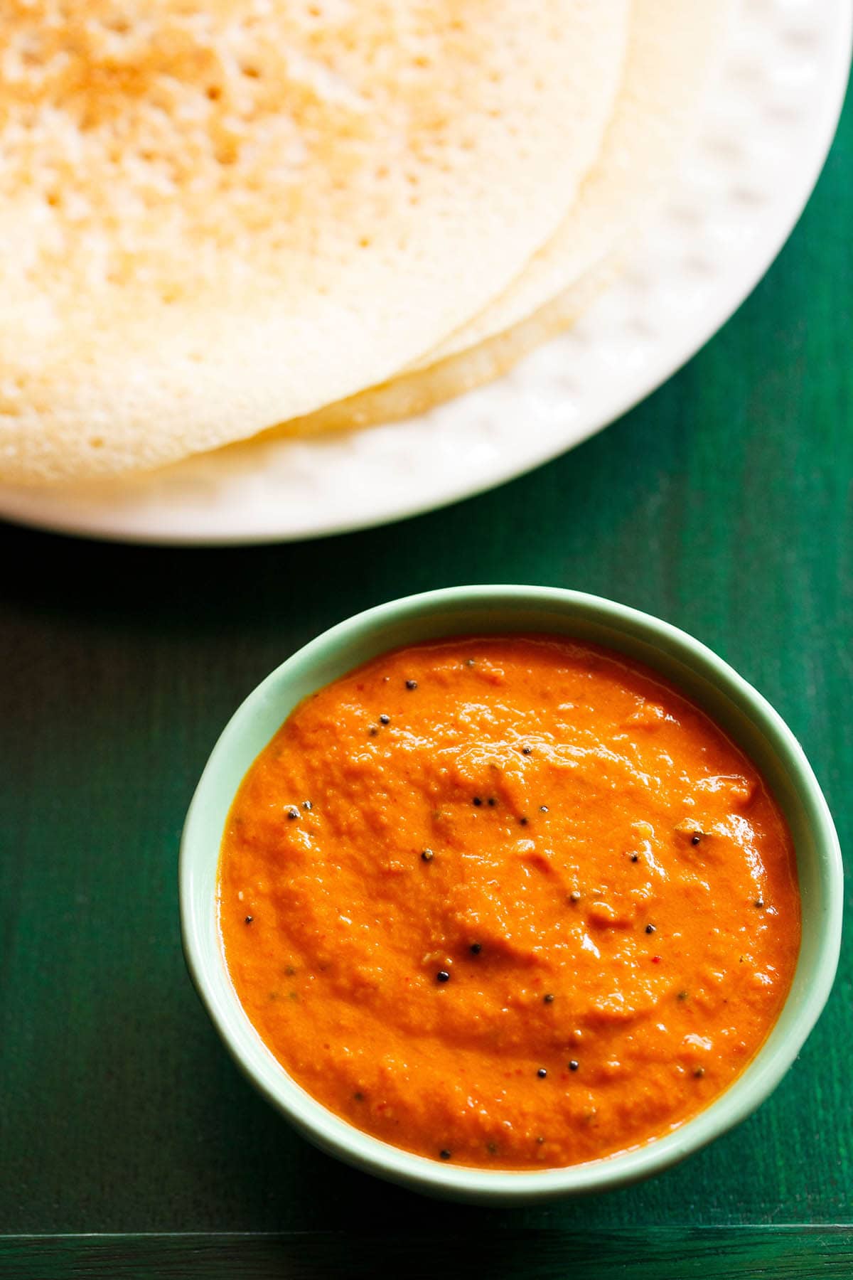 onion chutney in a green bowl with dosa stacked on a white plate on a dark green board