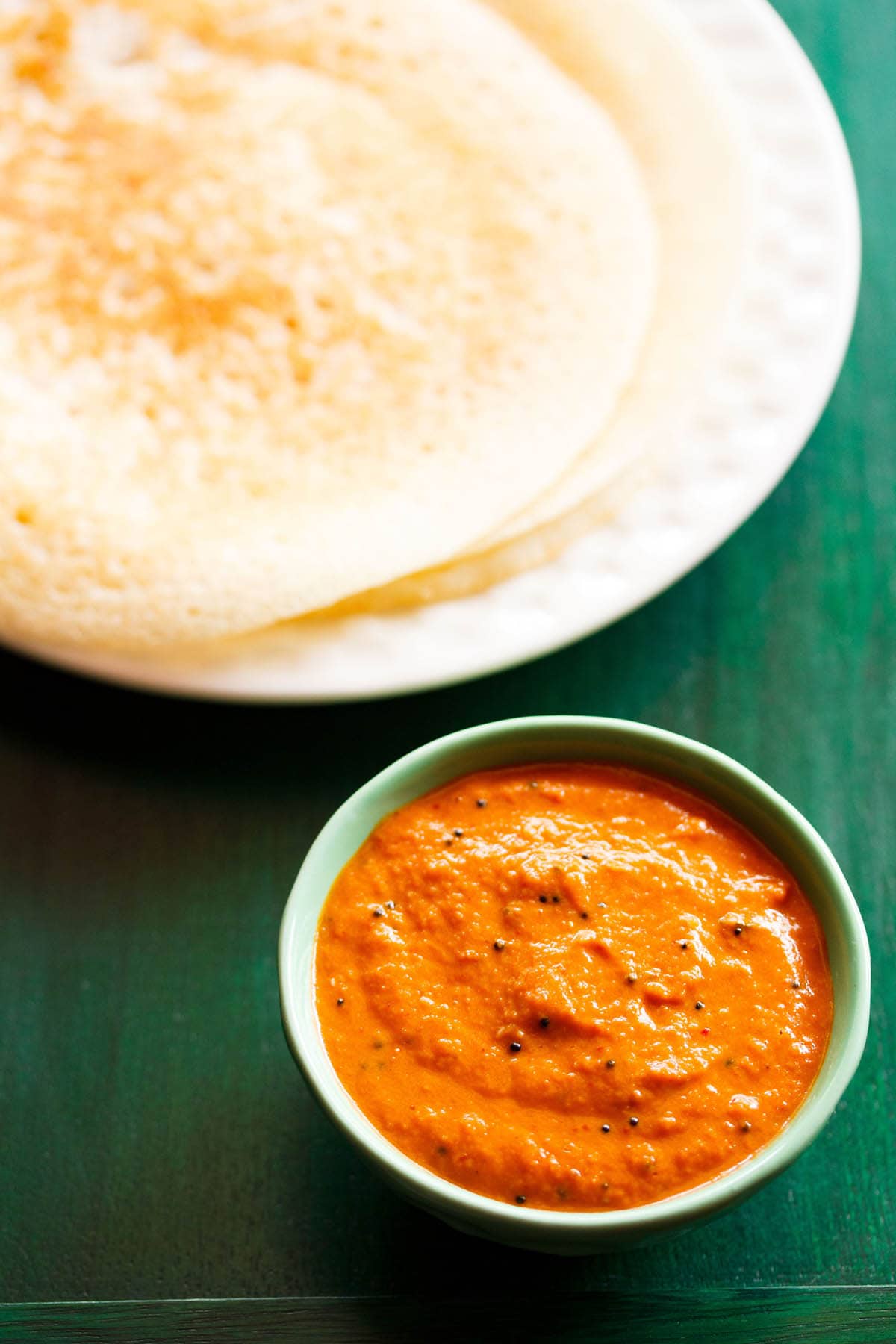 onion chutney in a green bowl with dosa stacked on a white plate on a dark green board 