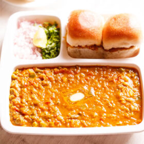 pav bhaji served in a rectangular serving tray with buttered pav and chopped onions, cilantro and lemon wedges on a white table