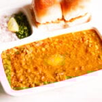 pav bhaji served in a rectangular serving tray with buttered pav and chopped onions, cilantro and lemon wedges on a white table