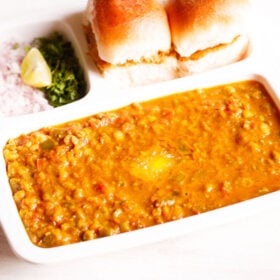 pav bhaji served in a rectangular serving tray with buttered pav and chopped onions, cilantro and lemon wedges on a white table