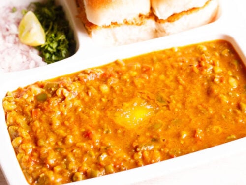 pav bhaji served in a rectangular serving tray with buttered pav and chopped onions, cilantro and lemon wedges on a white table