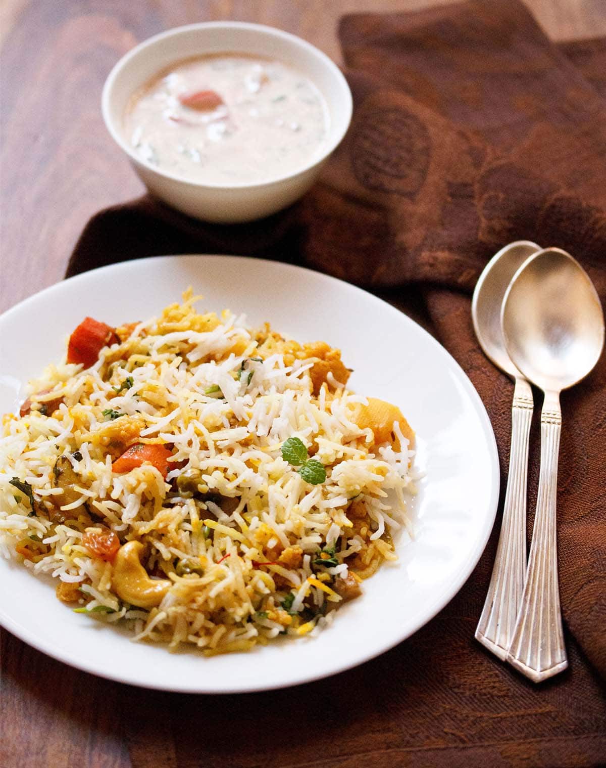 Top shot of veg dum biryani on white plate next to sauce in small white bowl and two spoons on wooden table