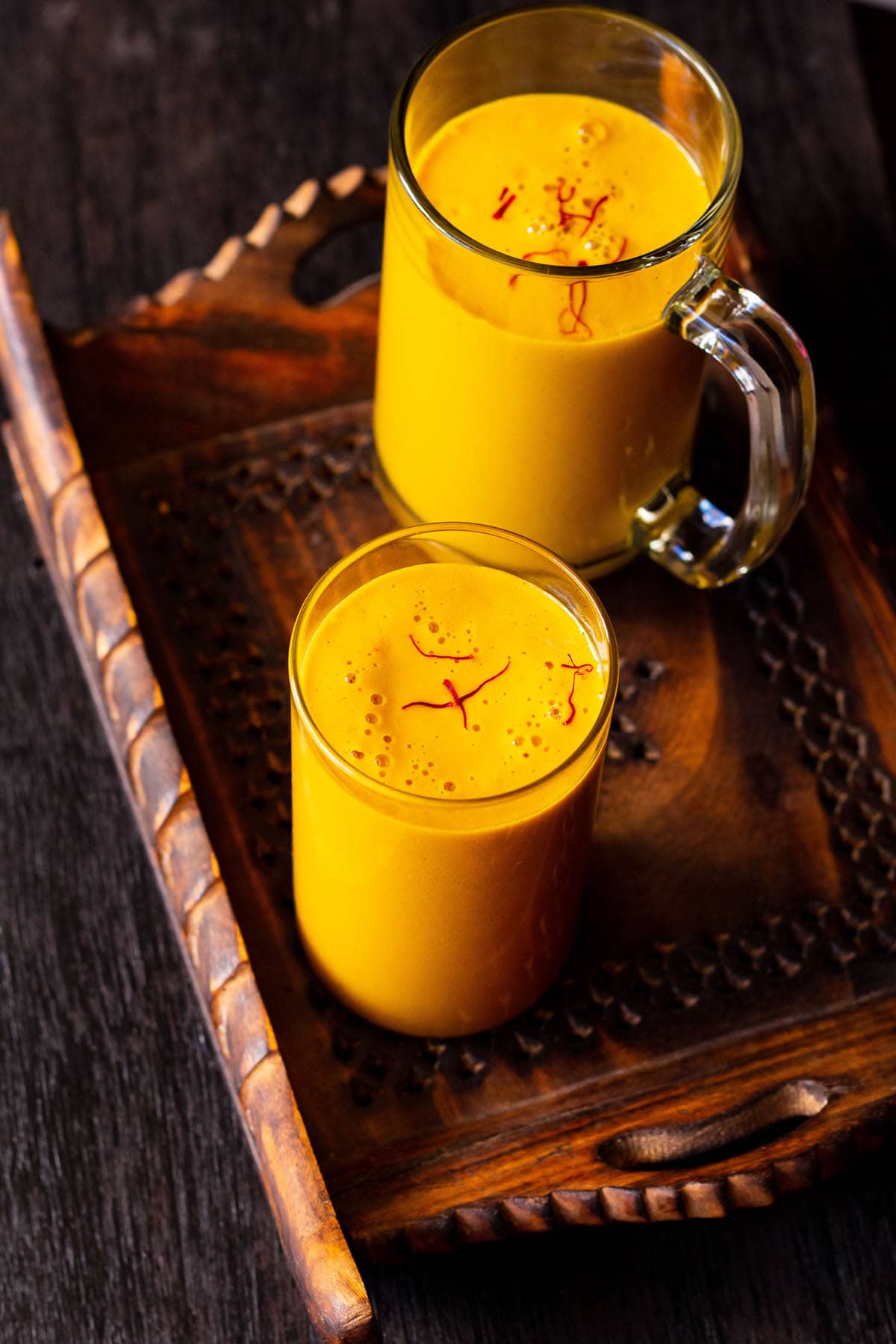 vegan mango milkshake in glass and a mug sprinkled with few saffron strands on a dark brown wooden tray