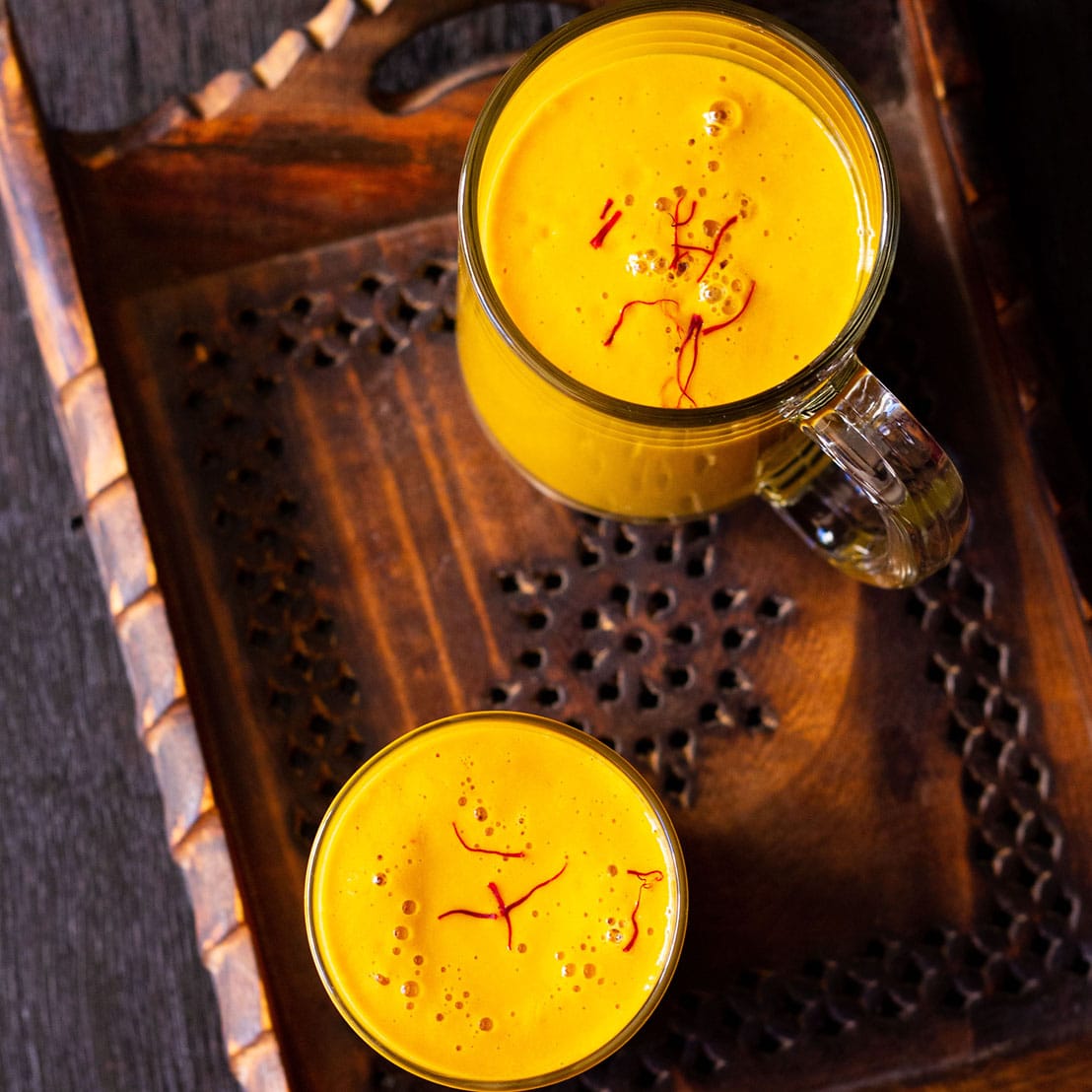 overhead shot of vegan mango shake in glass and a mug sprinkled with few saffron strands on a dark brown wooden tray