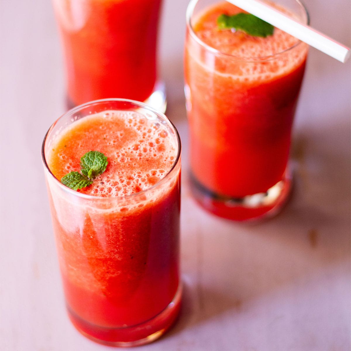 three tall clear glasses of watermelon orange juice garnished with sprigs of mint and orange segments on a white table