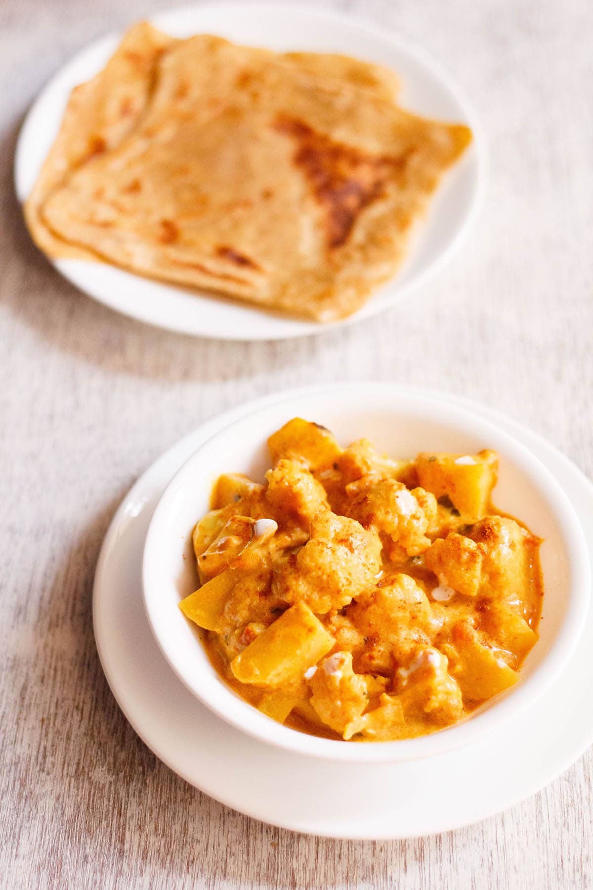 restaurant style aloo gobi in a white bowl with paratha in another plate on a white board