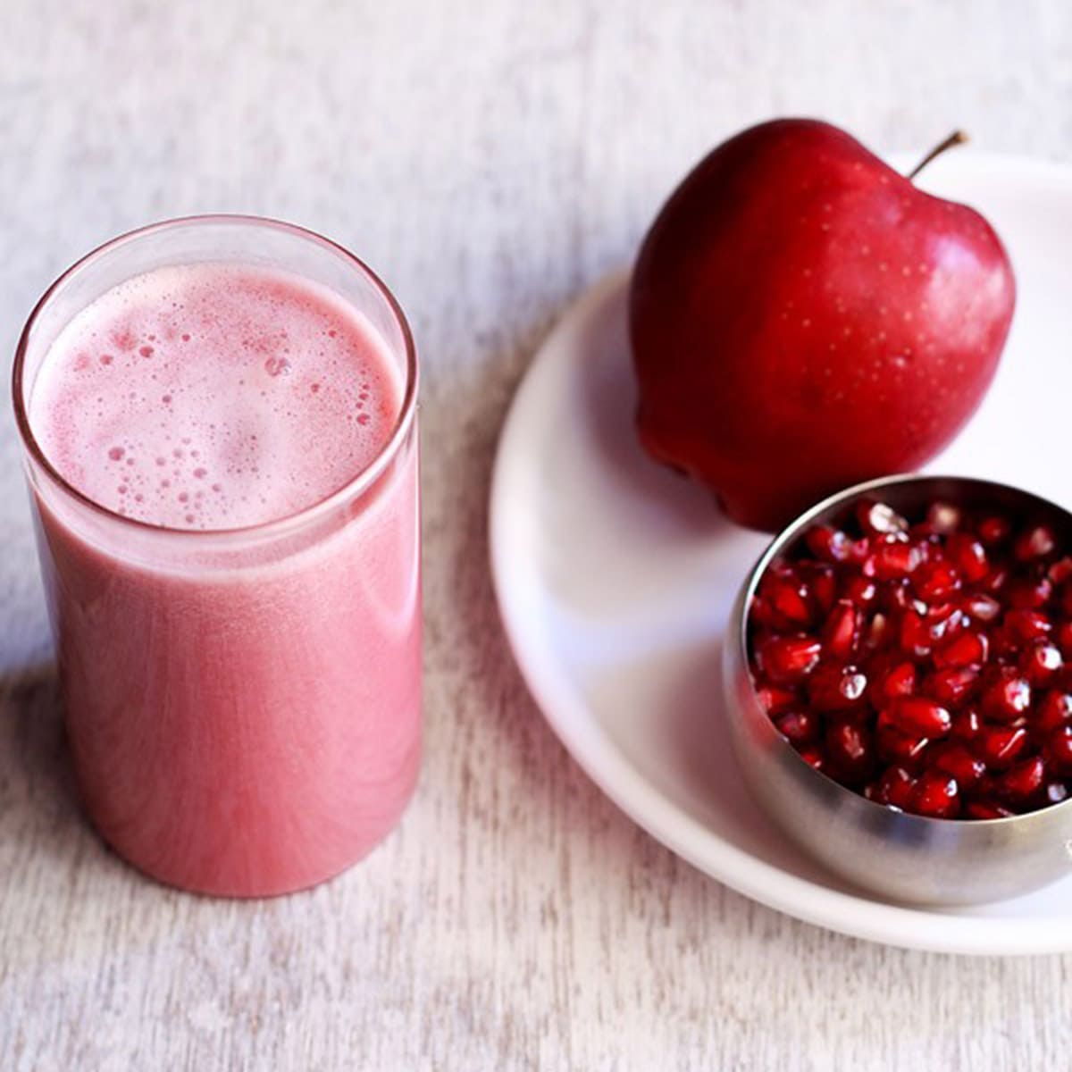 glass of apple pomegranate juice next to a silver bowl of pomegranate arils and a red apple on a white plate