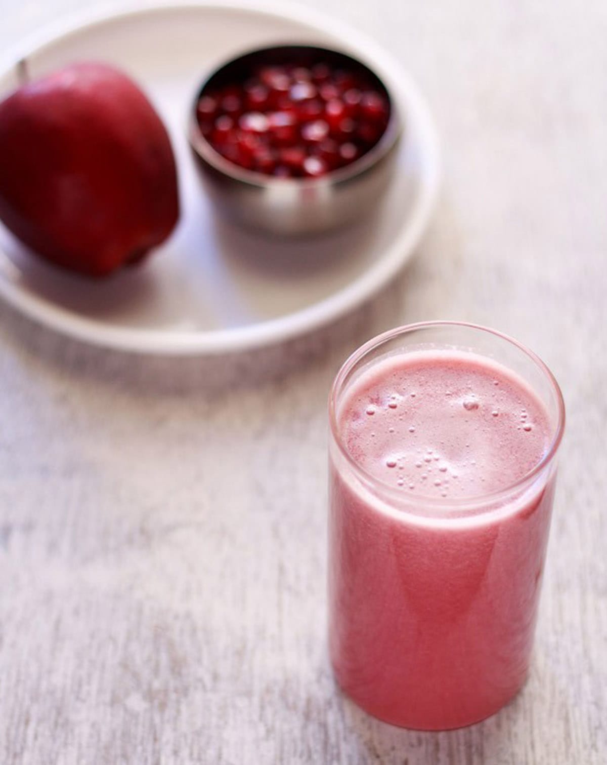 glass of apple pomegranate juice on a white-grey table with a white plate with an apple and pomegranate on it in the background