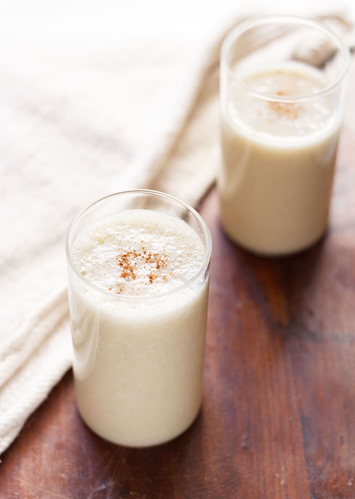 banana milkshake in two glasses with a light sprinkle of ground cinnamon on a dark brown table with a cream jute napkin by side