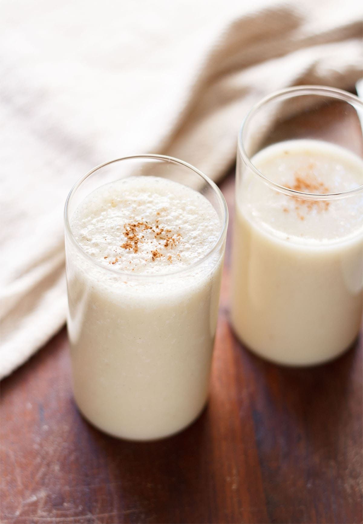 banana milkshake in two glasses with a light sprinkle of ground cinnamon on a dark brown table with a cream jute napkin by side