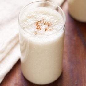 banana milkshake in a glass with a light sprinkle of ground cinnamon on a dark brown table with a cream jute napkin by side