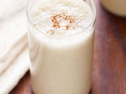 banana milkshake in a glass with a light sprinkle of ground cinnamon on a dark brown table with a cream jute napkin by side