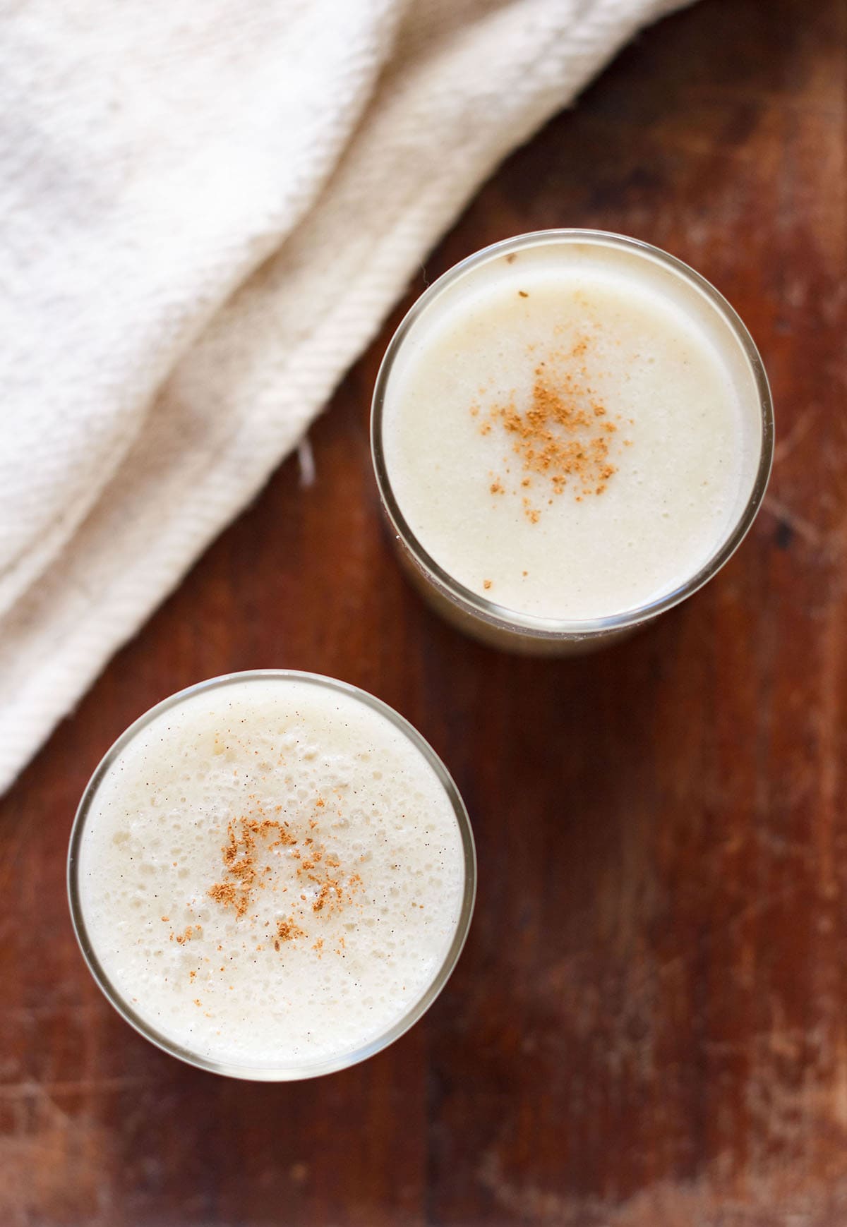 overhead shot of banana milkshake in two glasses with a light sprinkle of ground cinnamon on a dark brown table