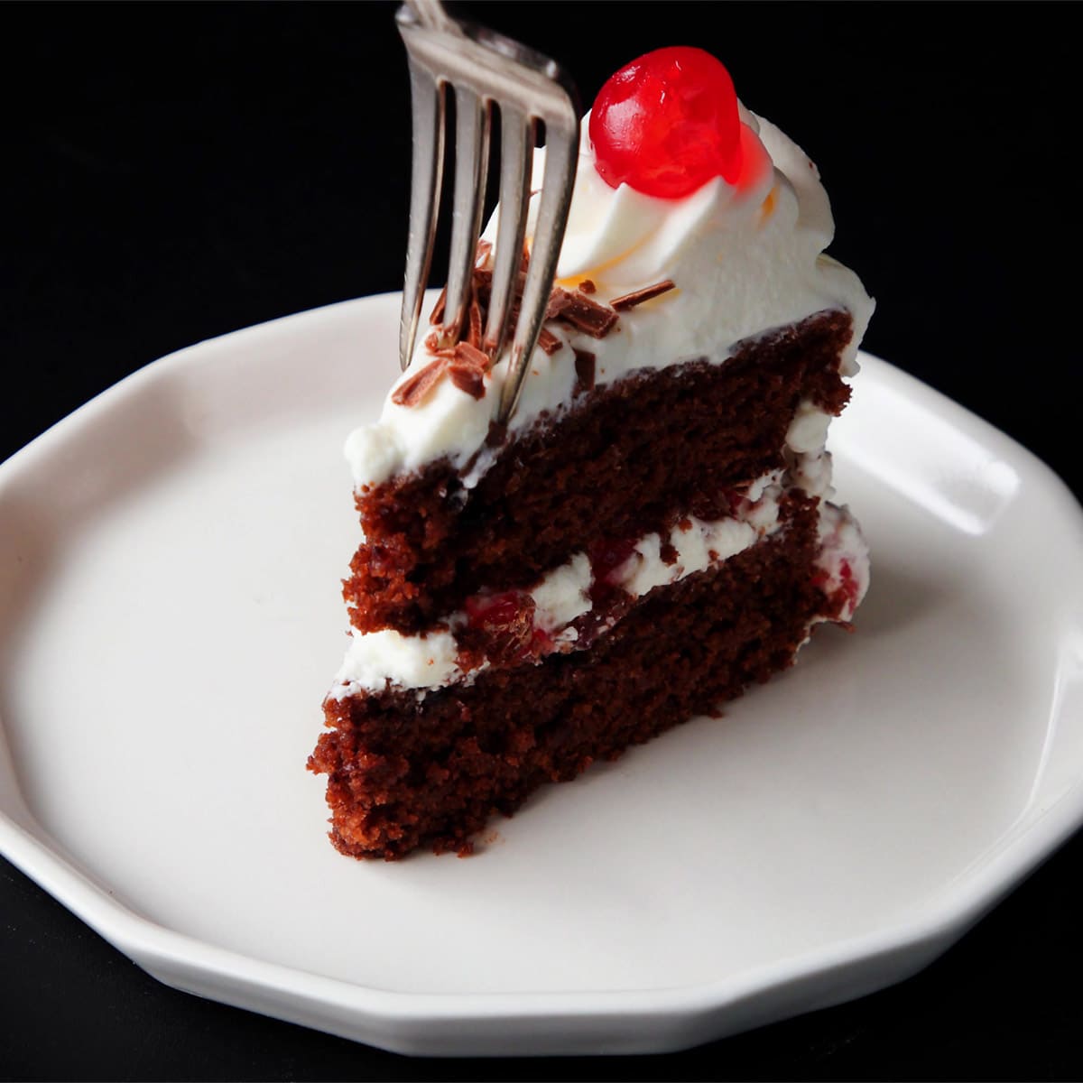 one slice of black forest cake on a white plate on a black background with a fork taking a bite