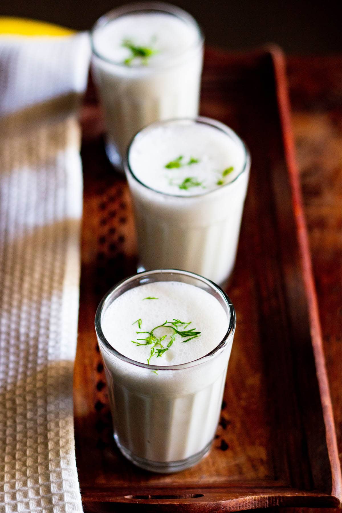 small three glasses filled with chaas placed on a wooden tray next to a white linen