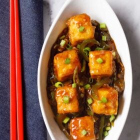 chilli paneer in oval white plate with red chopsticks on a blue fabric.