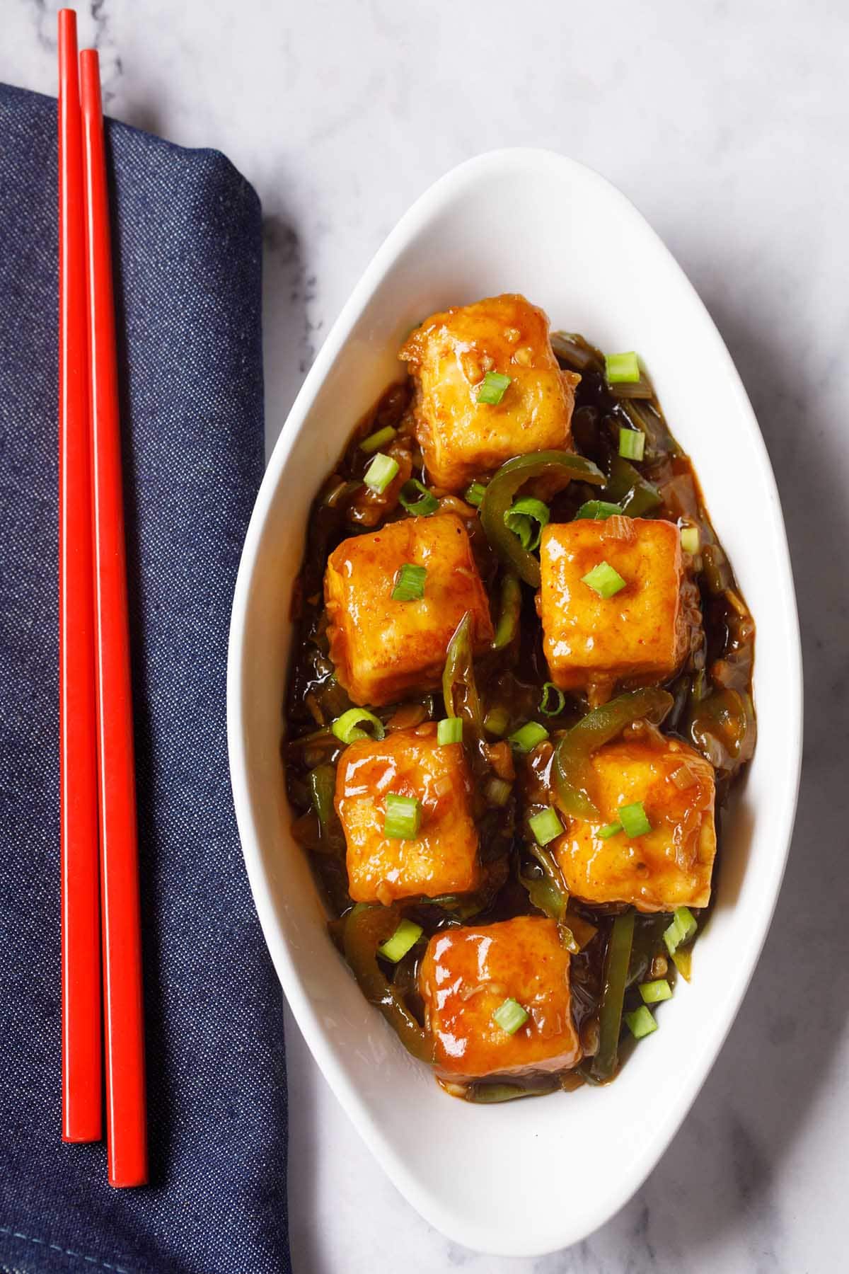 chilli paneer in oval white plate with red chopsticks on a blue fabric.