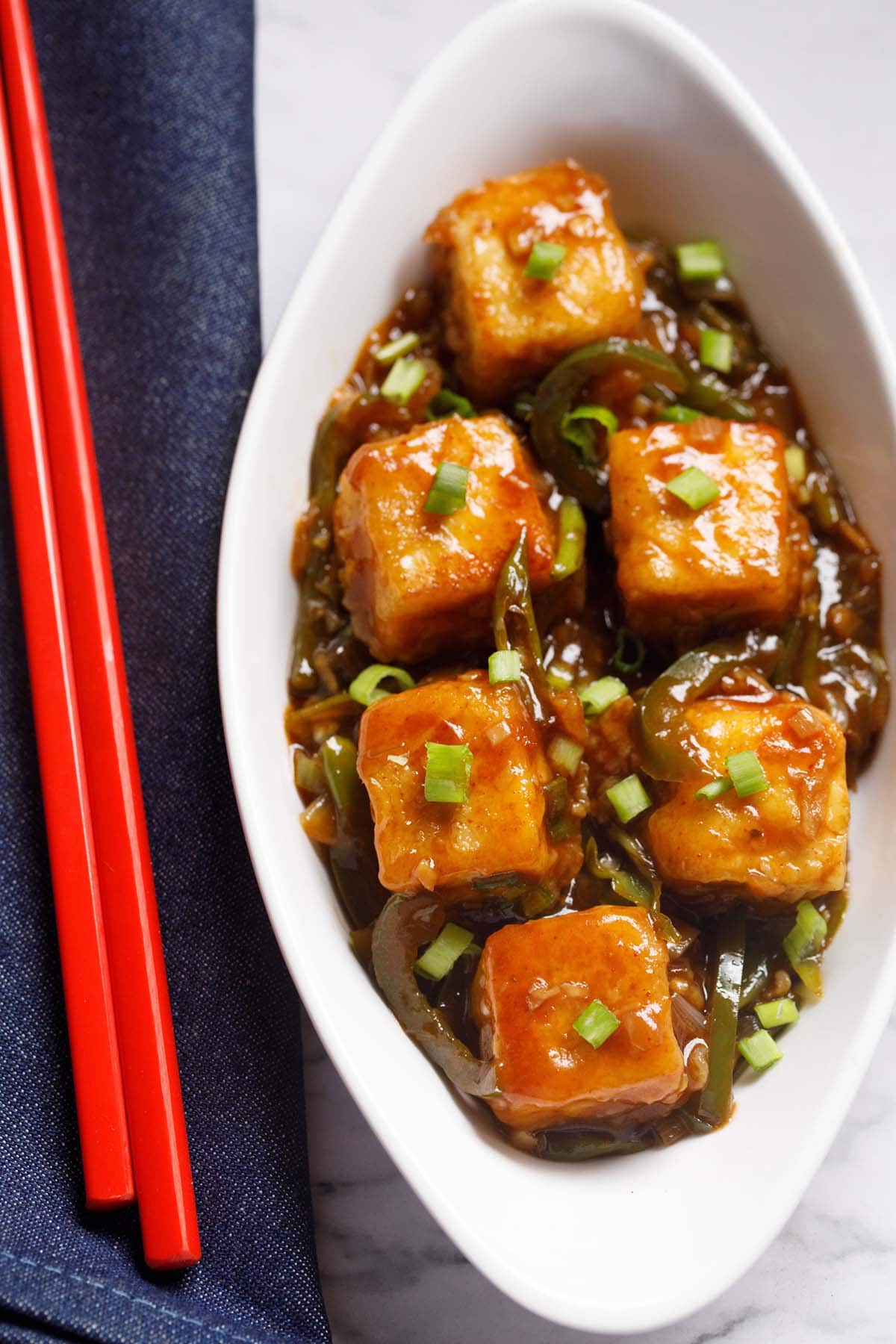 chilli paneer in oval white plate with red chopsticks on a blue fabric.