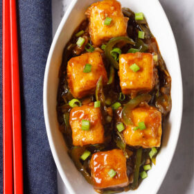 chilli paneer in oval white plate with red chopsticks on a blue fabric.