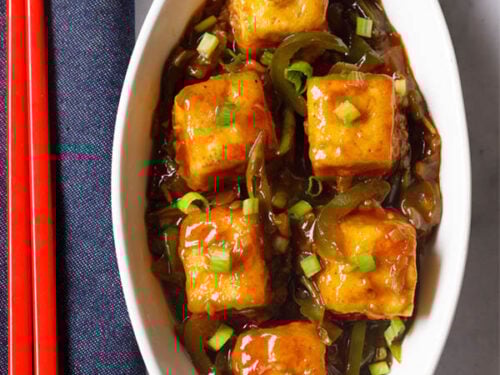 chilli paneer in oval white plate with red chopsticks on a blue fabric.