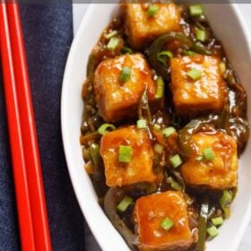 chilli paneer in oval white plate with red chopsticks on a blue fabric.