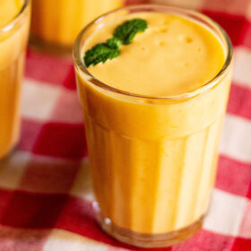 closeup shot of mango lassi with mint sprigs filled in a glass on a red and white checkered cloth