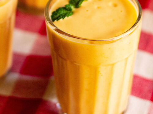 closeup shot of mango lassi with mint sprigs filled in a glass on a red and white checkered cloth