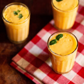 Mango lassi filled with sprigs of mint in three glasses on a red and white checkered napkin