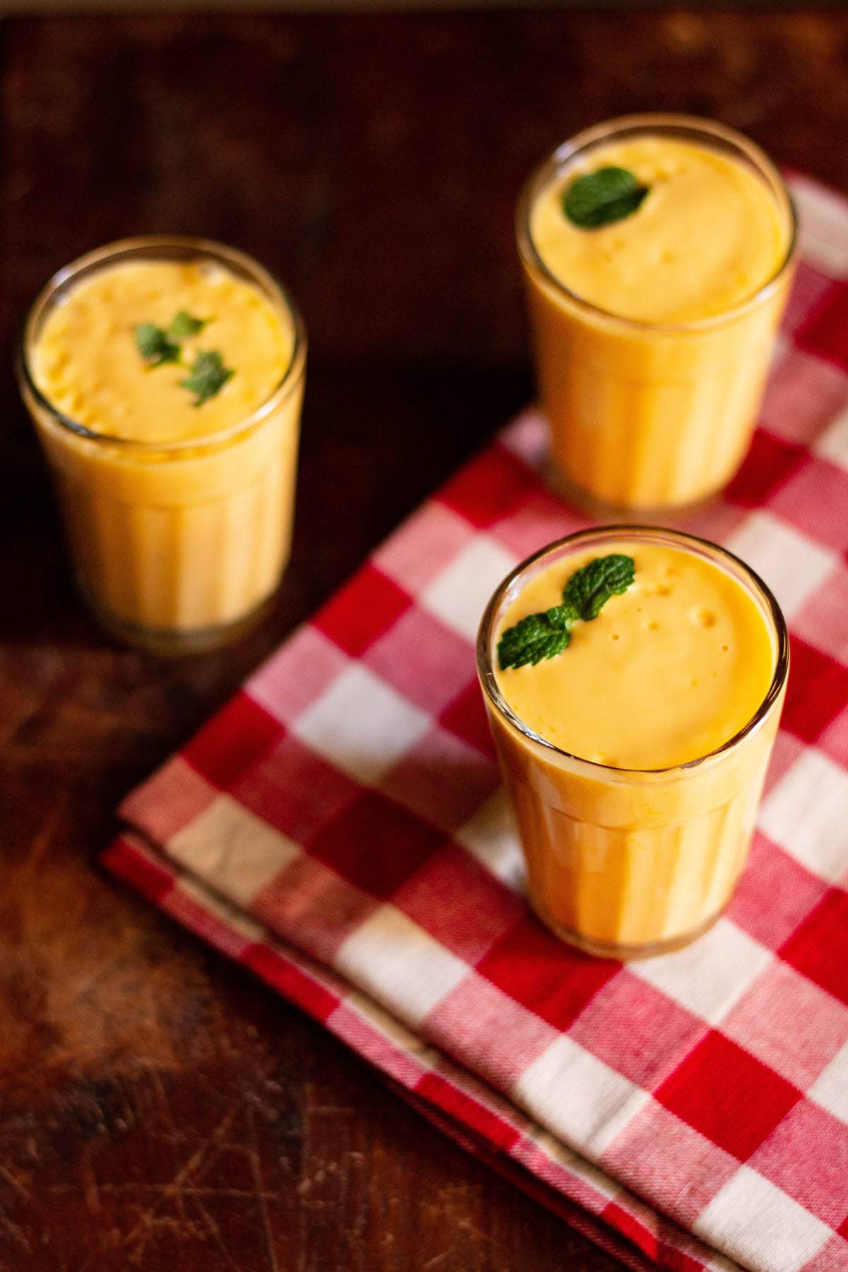 mango lassi with mint sprigs filled in three glasses on a red and white checkered napkin