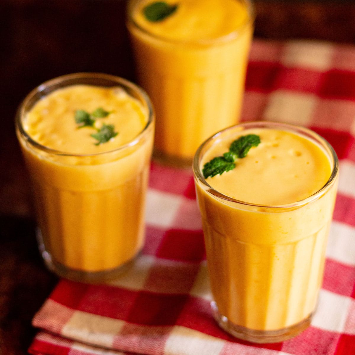 mango lassi with mint sprigs filled in three glasses on a red and white checkered napkin