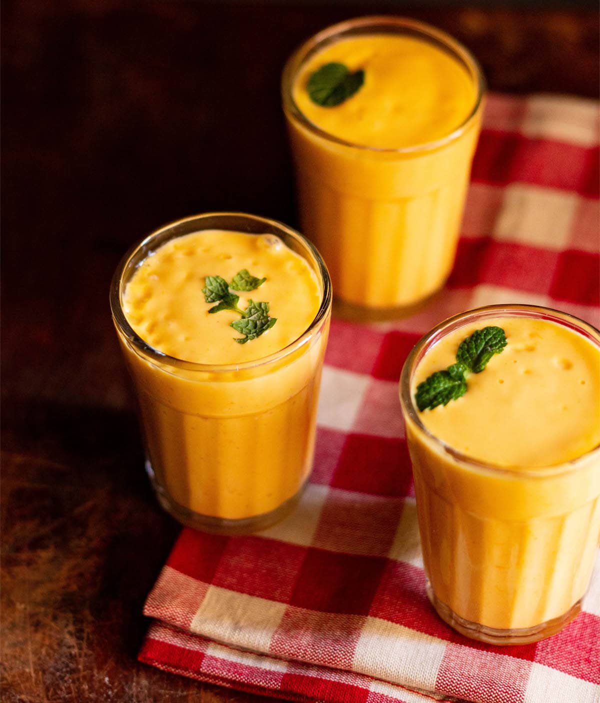 mango lassi with mint sprigs filled in three glasses on a red and white checkered napkin