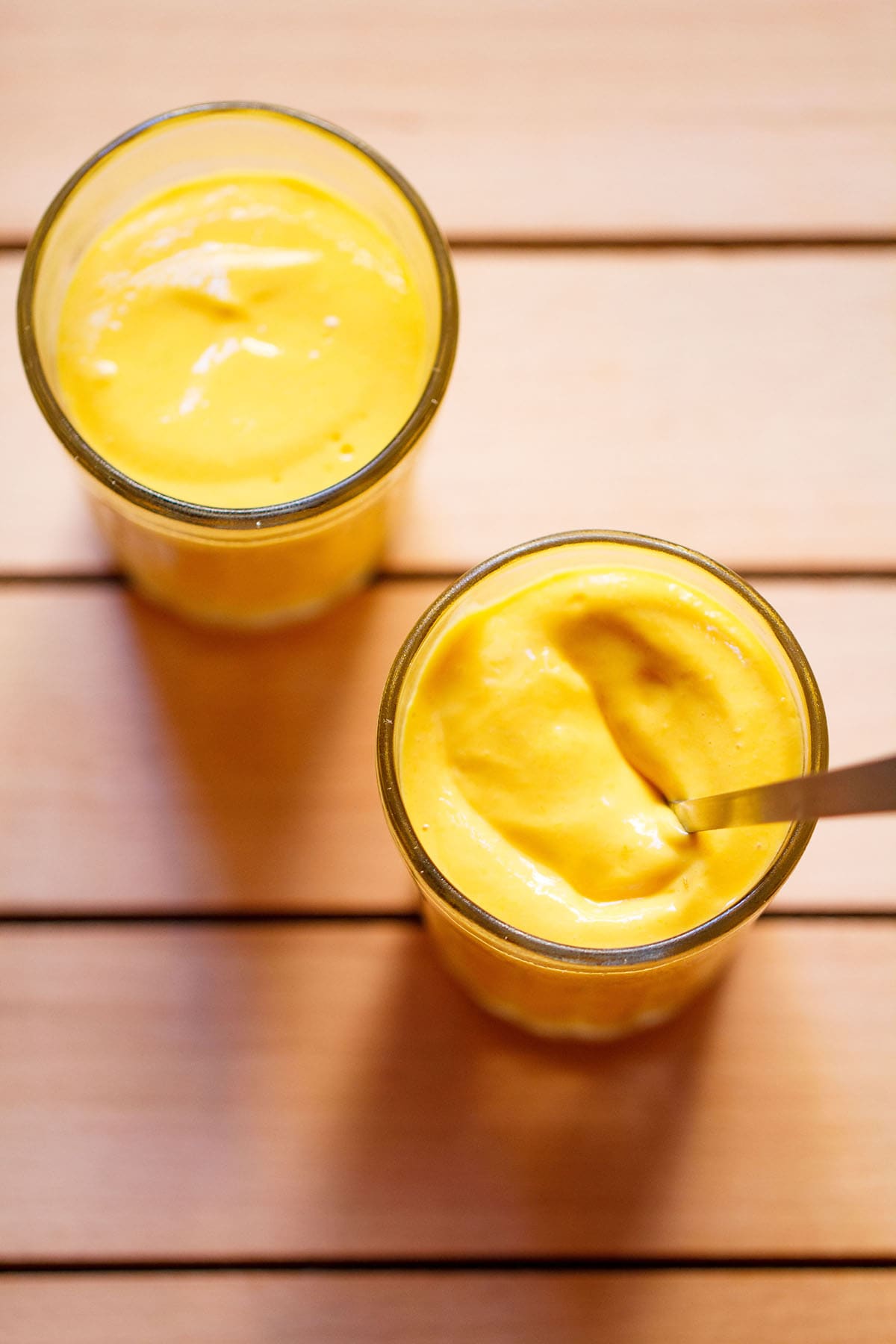 mango mousse in two shot glasses with a spoon inside on glass on a dark beige wooden board