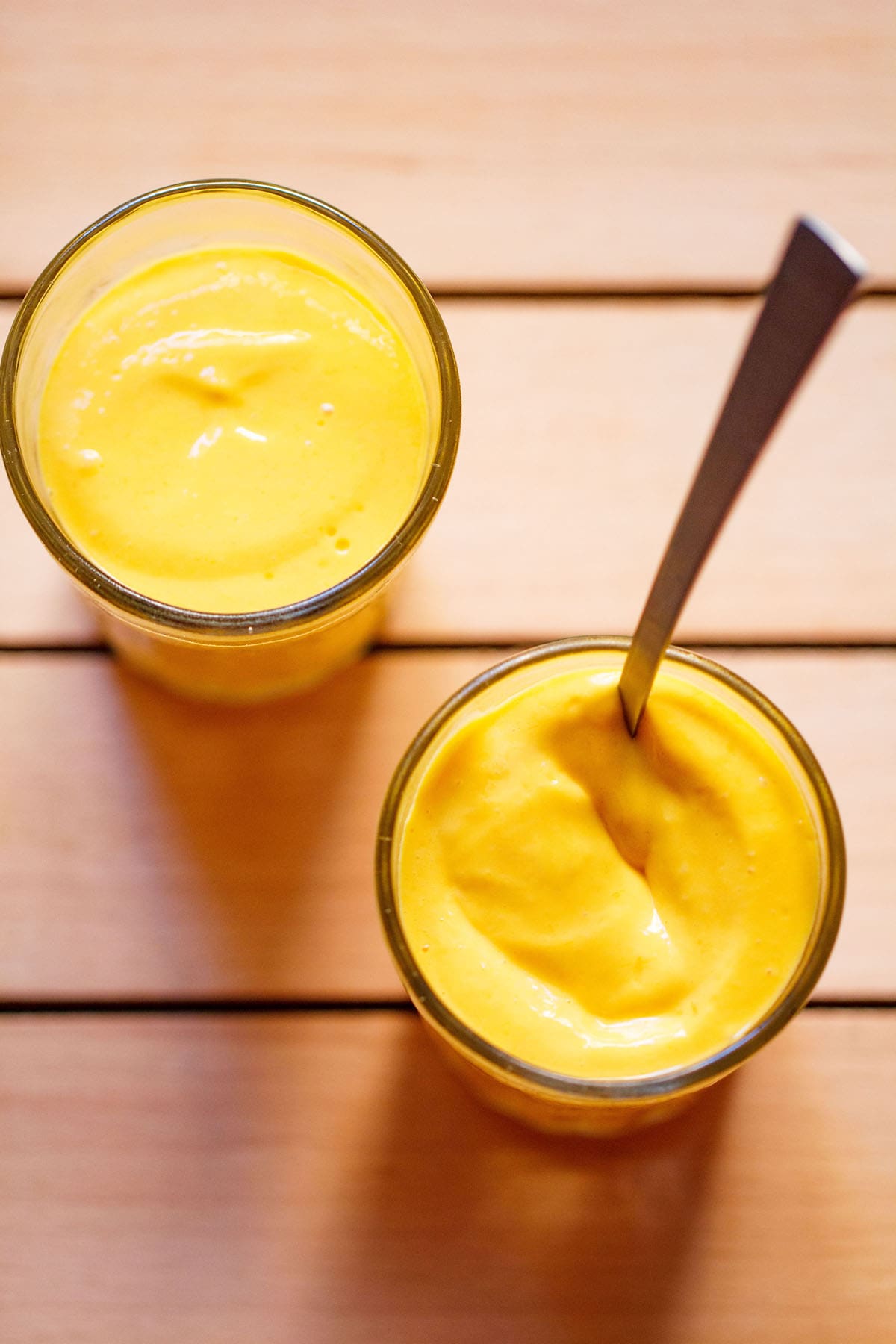 mango mousse in two shot glasses with a spoon inside on glass on a dark beige wooden board