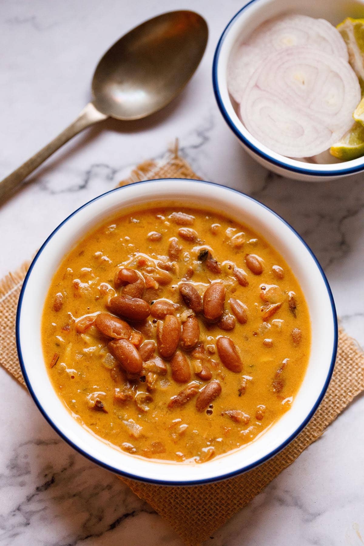 rajma masala in a blue rimmed white bowl on a jute cloth with a brass spoon and bowl with onion rounds and lemon wedges