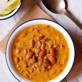 overhead shot of rajma recipe in a blue rimmed white bowl on a jute cloth with a brass spoon and bowl with onion rounds and lemon wedges