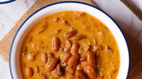 overhead shot of rajma recipe in a blue rimmed white bowl on a jute cloth with a brass spoon and bowl with onion rounds and lemon wedges