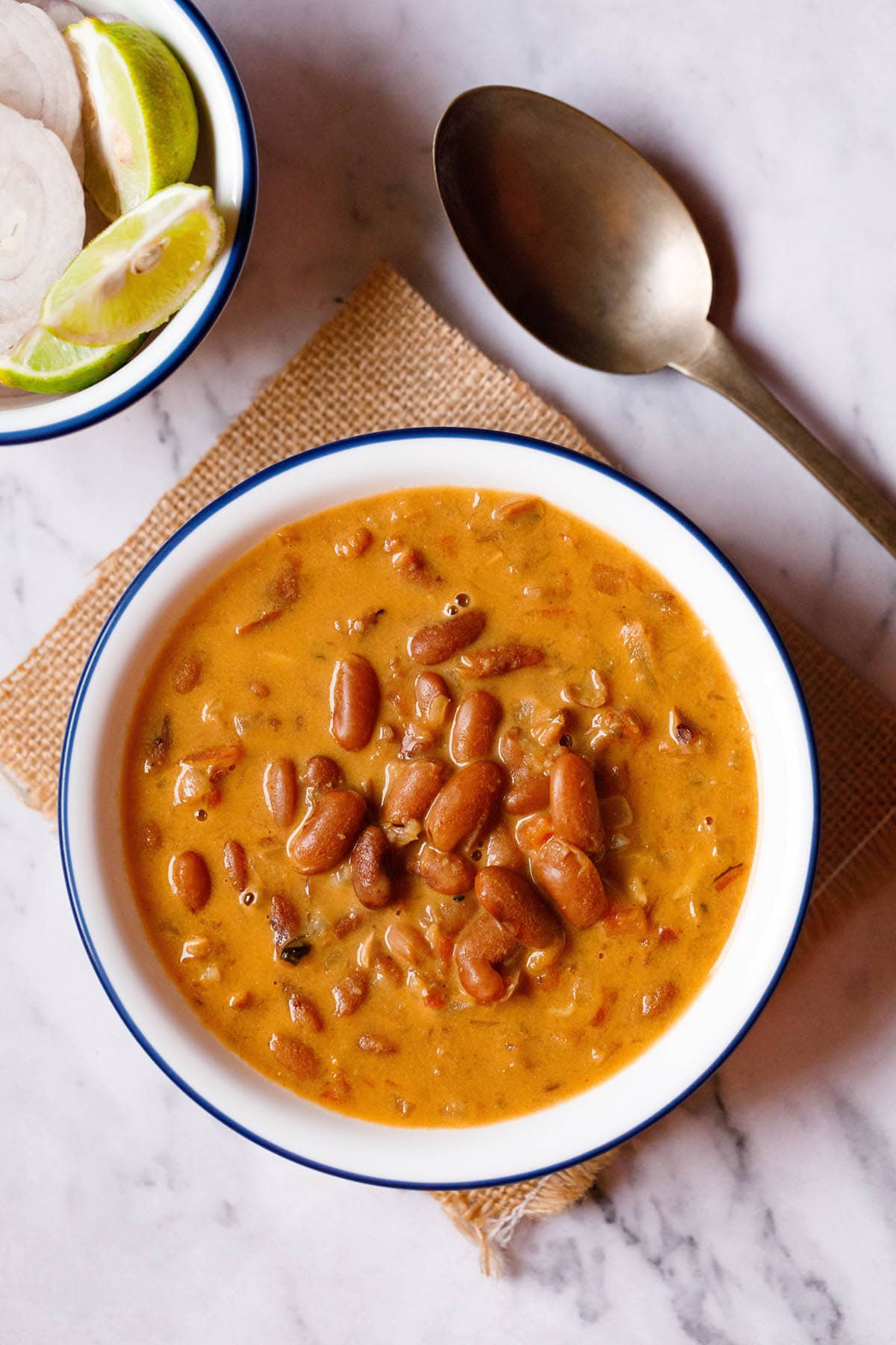 overhead shot of rajma recipe in a blue rimmed white bowl on a jute cloth with a brass spoon and bowl with onion rounds and lemon wedges