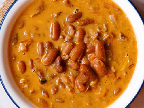 overhead shot of rajma recipe in a blue rimmed white bowl on a jute cloth with a brass spoon and bowl with onion rounds and lemon wedges